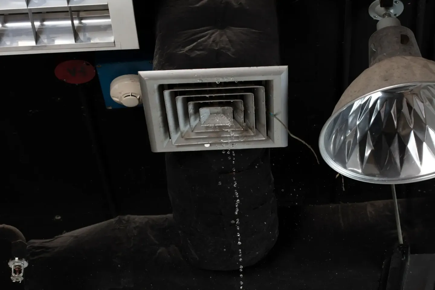 Light fixture sitting atop a black ceiling, with a ventilator and a light on the right side